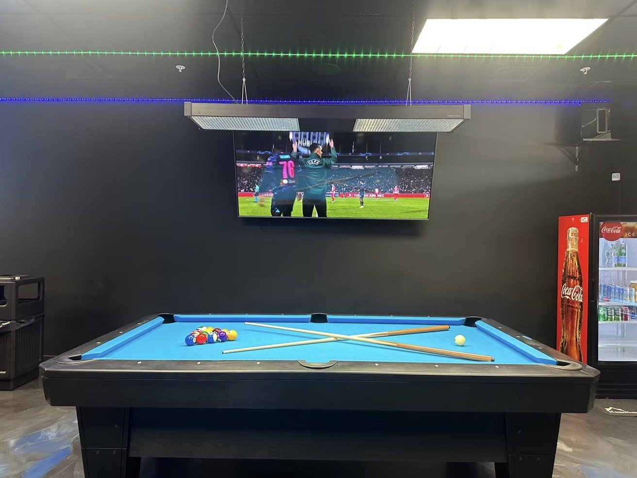 A Diamond table beneath a big screen showing the match, with a drinks fridge beside it